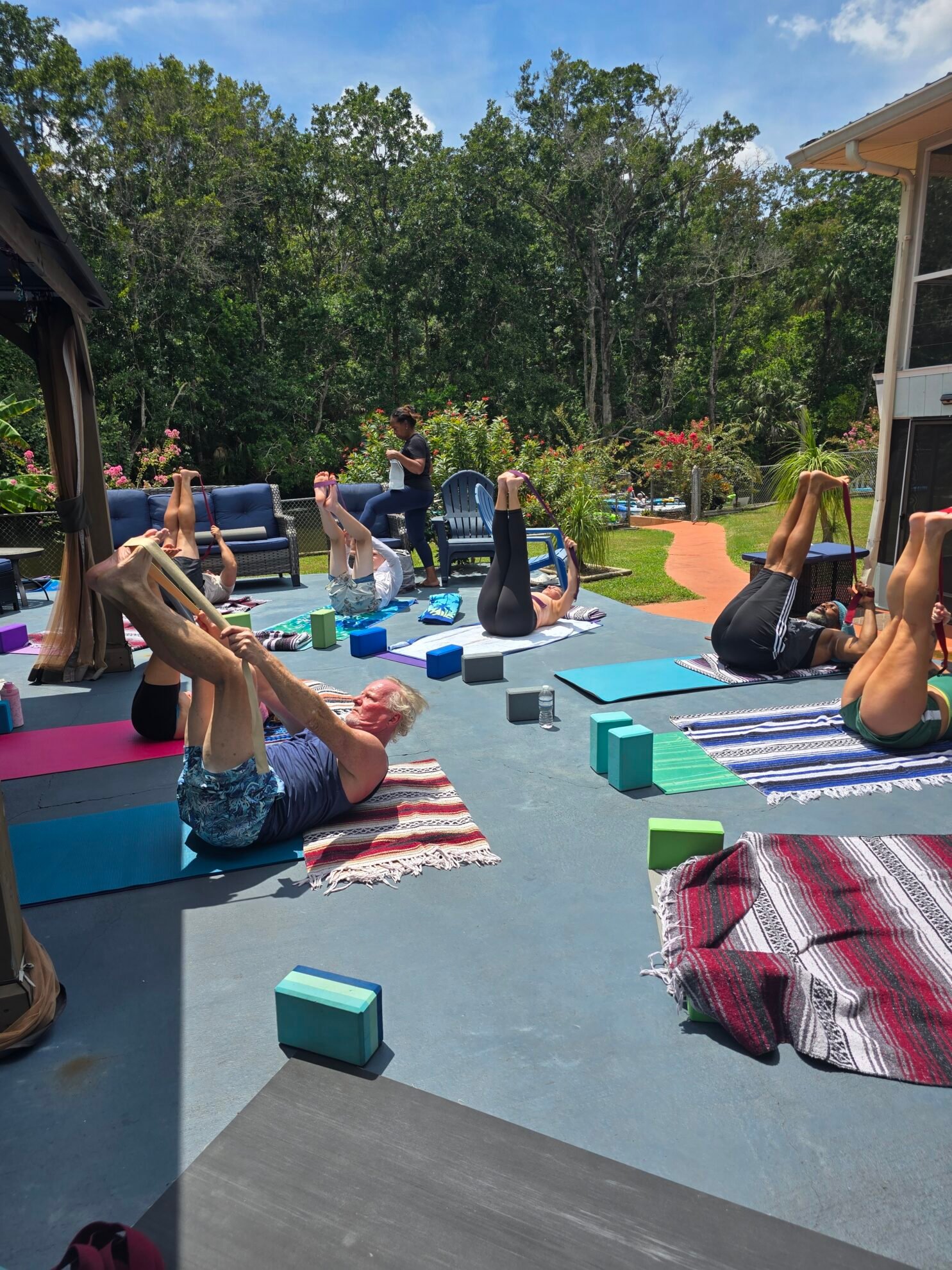 Students lying on their backs stretching their legs overhead with yoga straps during an outdoor Sacred Space Yoga class