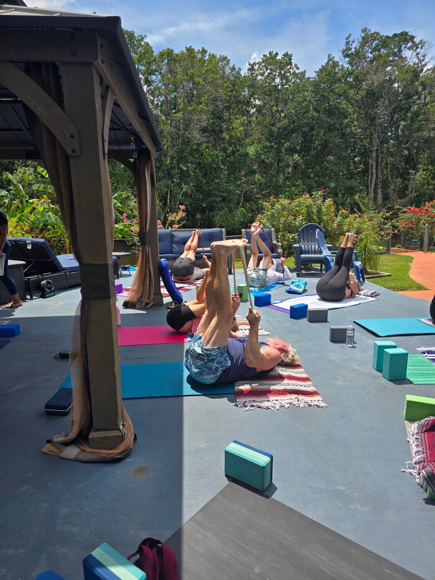 Older man using a yoga strap for a reclined hamstring stretch during an outdoor Sacred Space Yoga class on a sunny patio
