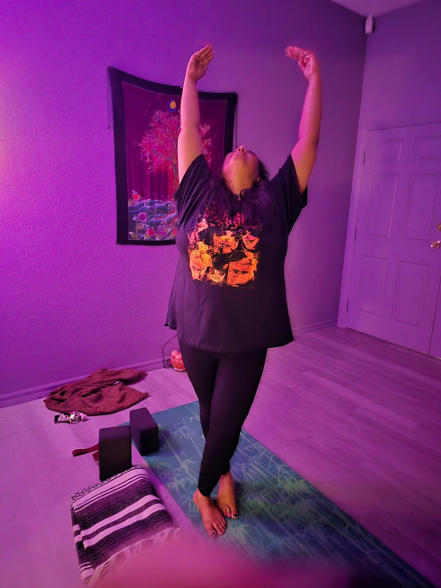 Woman reaching both arms overhead in a standing backbend on a yoga mat in a purple-lit yoga room at Sacred Space Yoga