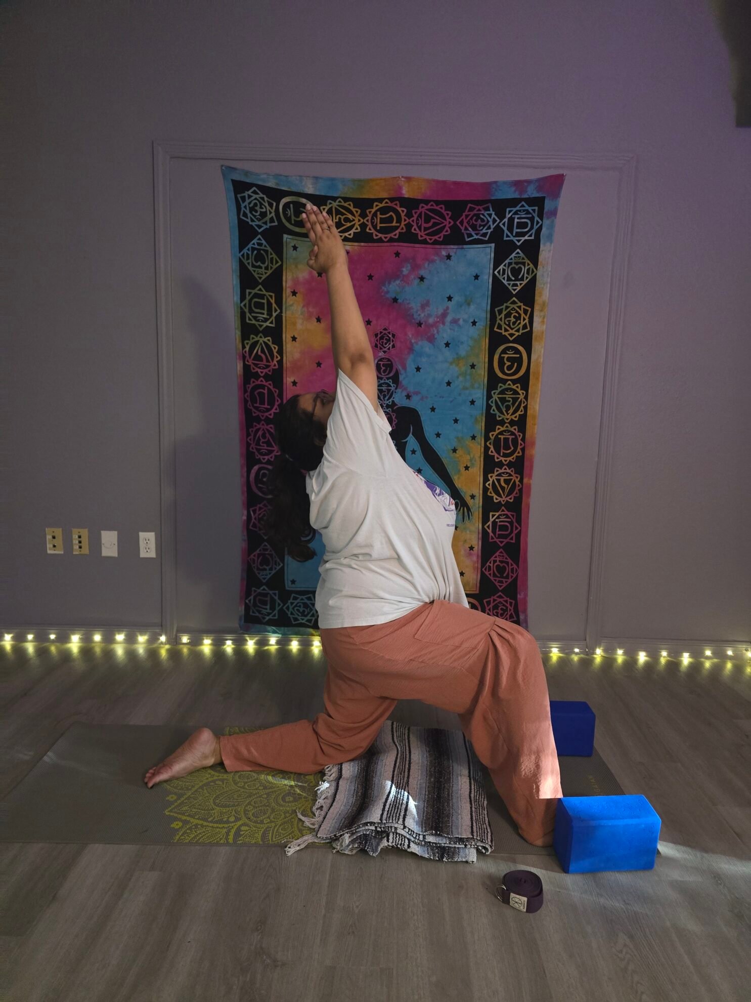 Woman in a crescent low lunge with one arm raised and her knee on a folded blanket in front of a chakra tapestry at Sacred Space Yoga